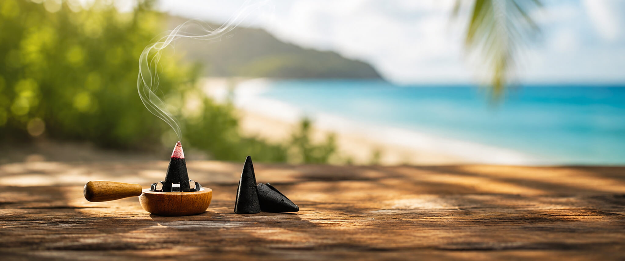 A small incense burner with a smoking incense cone and a black cone resting beside it, placed on a wooden surface with a blurred tropical beach and ocean background.