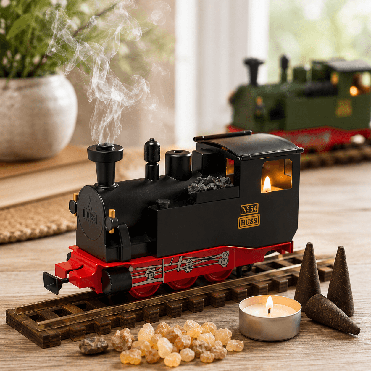 HUSS Incense Train black, locomotive incense burner with smoke rising from the chimney, a glowing tealight, and incense cones on railway tracks on a table with another train in green in the background.