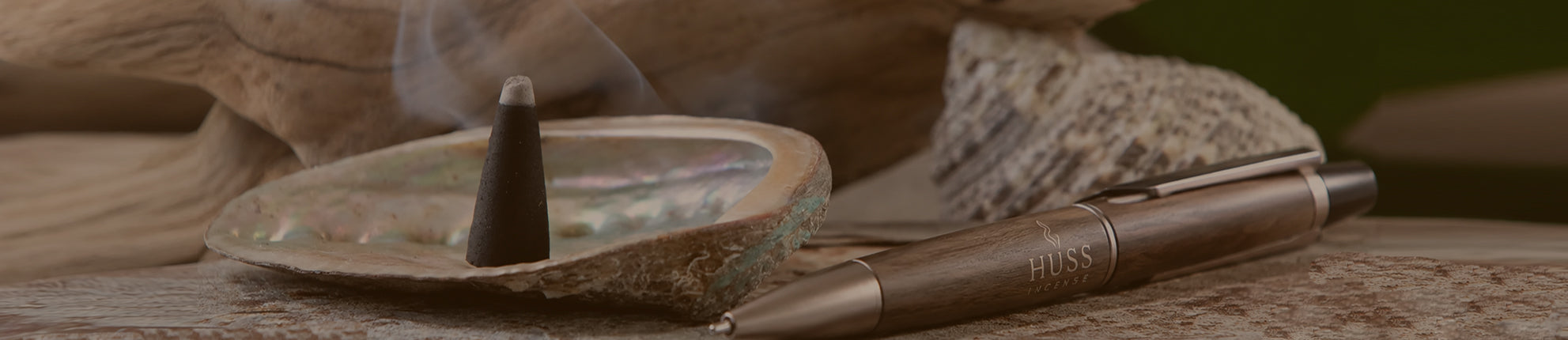 A close-up of a metallic pen resting on a textured surface next to a small incense burner with a smoking incense cone, with driftwood and rocks in the background.