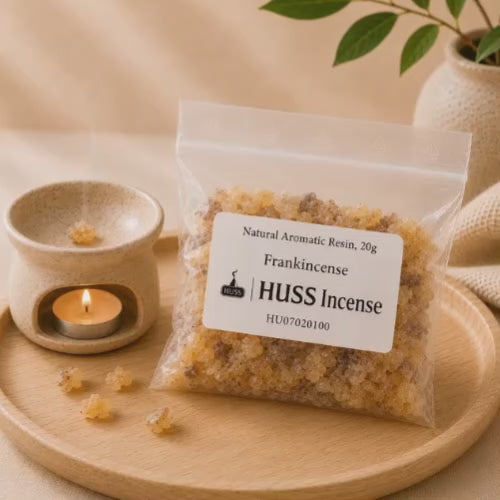 Video of a clear plastic bag of frankincense aromatic resin sits on a round wooden tray beside a small lit candle holder with a burning tealight. The label reads 'Natural Aromatic Resin, 20g Frankincense | HUSS Incense'.