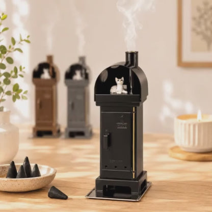 Video of a HUSS Incense woodburner black with a cat steaming out of the chimney displayed on a wooden table with two more ovens in the back.