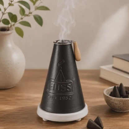 Video of a HUSS Incense Meiler black, incense burner with a burning incense cone and smoke rising, displayed on a wooden table in a cozy scene.
