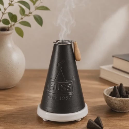 Video of a HUSS Incense Meiler black, incense burner with a burning incense cone and smoke rising, displayed on a wooden table in a cozy scene.
