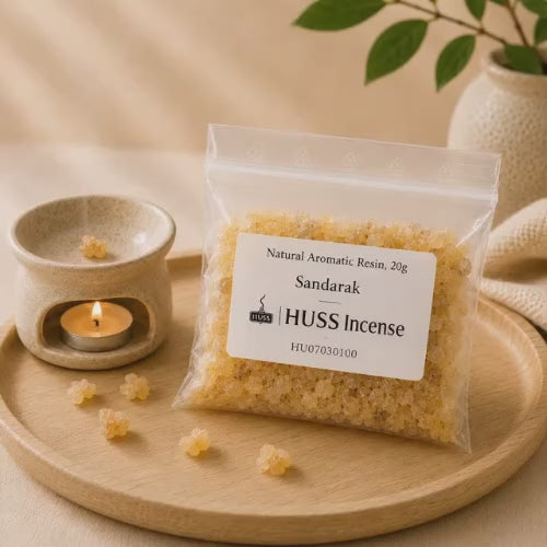 Video of a clear plastic bag of Huss Incense Sandarak natural aromatic resin sits on a round wooden tray beside a small lit candle holder with a few loose resin pieces scattered around. The label reads 'Natural Aromatic Resin, 20g Sandarak | HUSS Incense'.
